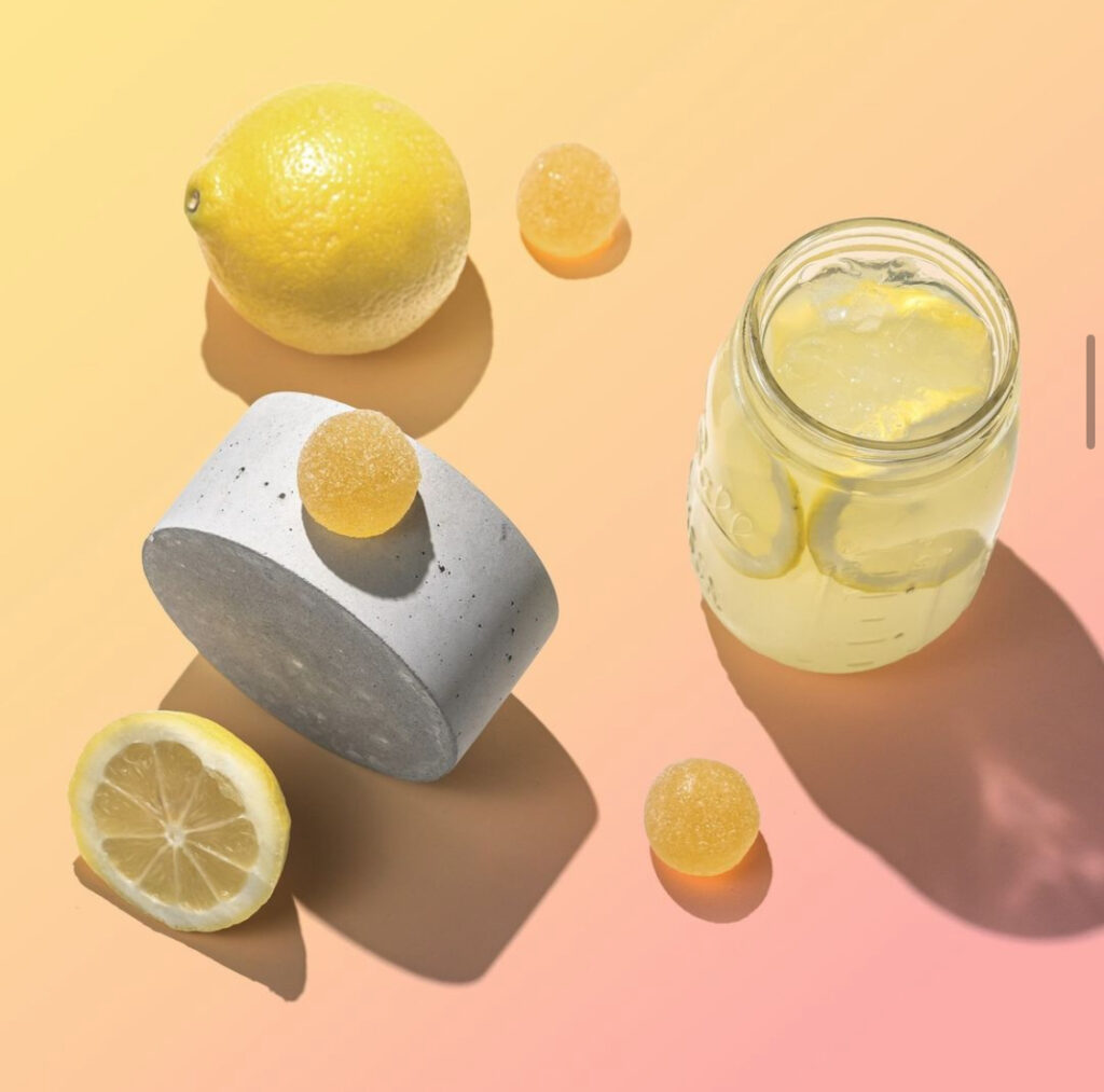 Yellow cannabis gummies surrounded by a glass of lemonade and lemons on a pink and yellow background.