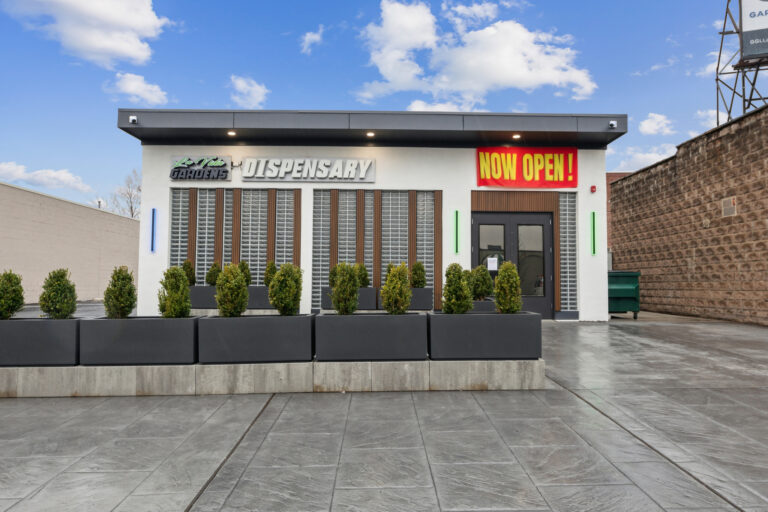 Exterior of white building with La Vida Gardens sign and a red banner with yellow font that reads Now Open.