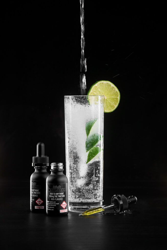 Bottle of ILLICIT brand cannabis tincture next to an iced beverage with a lime wedge.