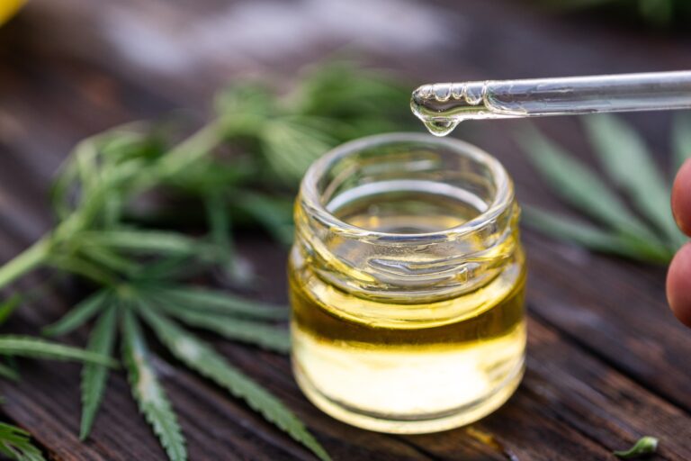 Small jar of cannabis oil with dropper on top of cannabis leaves.