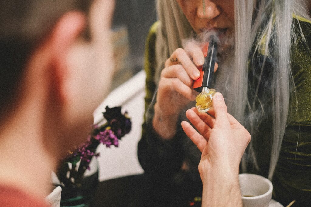 Man holds an open jar of a cannabis concentrate as a woman uses a nectar collector to consume the concentrate.