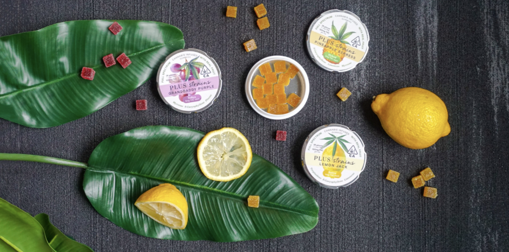 Flat lay of full-spectrum cannabis-infused gummy tins in multiple strain varieties with fresh lemons and tropical leaves, showing the range of THC edible products available for experienced users