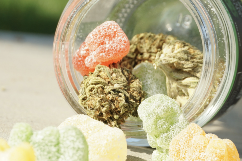 Assorted sugar-coated THC gummies in multiple flavors spilling from a glass jar alongside cannabis flower buds, showing the variety of edible options available at New Jersey dispensaries