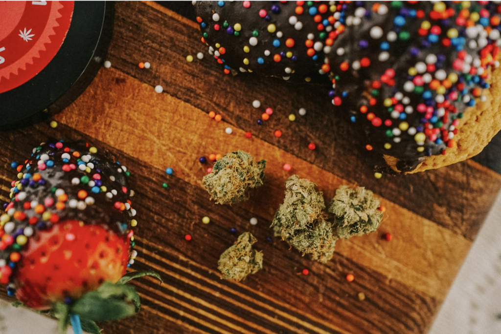 Cannabis flower buds alongside chocolate-sprinkle cookies and a strawberry on a wooden cutting board, representing the variety of THC edible formats available at NJ dispensaries