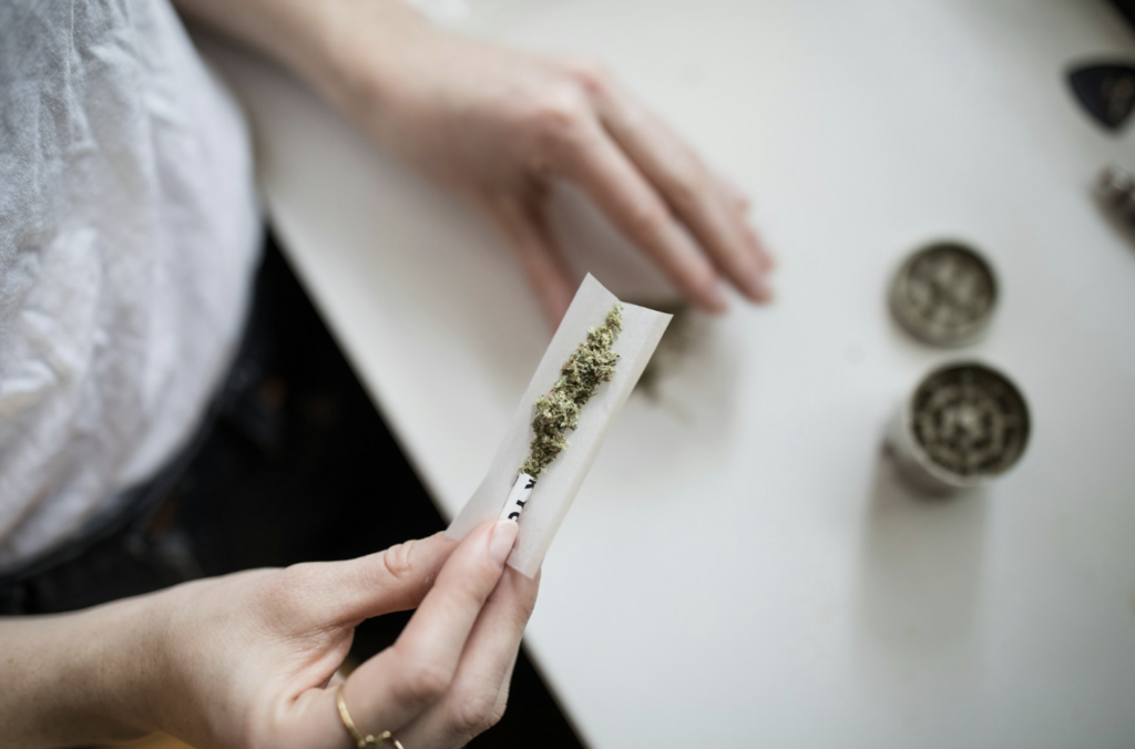 Source - Alt: Hands rolling a cannabis joint on a white surface with a metal grinder nearby, representing the experienced flower smokers who find a 10 mg edible dose moderate rather than strong