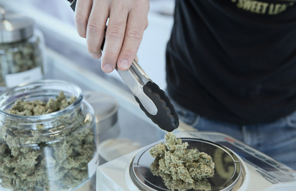 Dispensary budtender using tongs to weigh cannabis flower on a digital scale next to a jar of buds, illustrating how NJ dispensaries measure grams and eighths for each transaction