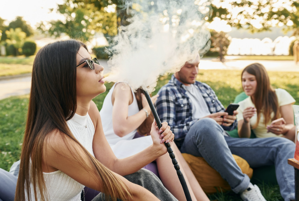 Woman exhaling smoke while sitting outdoors in a park with friends nearby, illustrating the type of public setting where cannabis consumption is illegal in NJ, with fines starting at $250