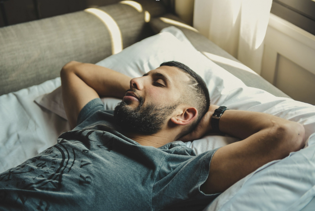 Man relaxing in bed with his hands behind his head and eyes closed, representing the restful sleep that myrcene-dominant cannabis strains are used to promote